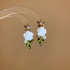 Close-up of elegant white rose earrings with green leaves, pearls, and rhinestones.