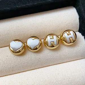 Close-up detail of gold-plated round stud earrings featuring H-letter and white heart inlays