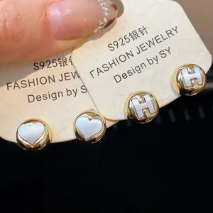 Close-up detail of round gold stud earrings featuring heart and H letter motifs.