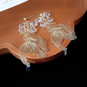 Close-up of gold filigree goldfish earrings with blue water-drop beads on a wooden tray.