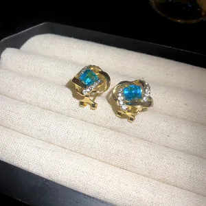 Detalle de lujosos pendientes dorados con piedra azul cuadrada y micro-pavé de cristales.