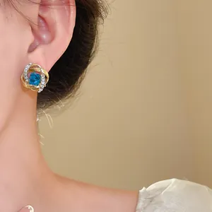 Primer plano de un pendiente de oro con cristal azul cuadrado usado en la oreja.