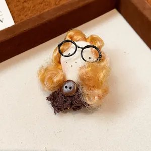 Close-up of a handmade doll brooch featuring blonde curls and black round glasses