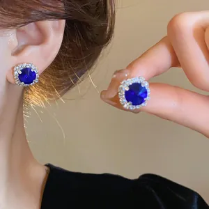 Close-up of royal blue round halo stud earrings worn on ear and held by hand