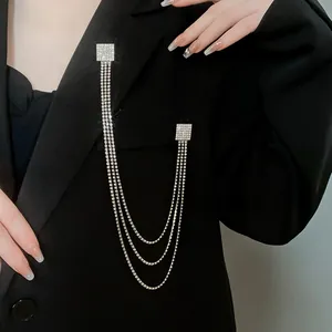 Dettaglio di una spilla quadrata con strass e nappe a catena su un blazer nero.