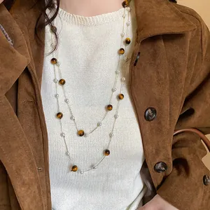 Collar largo de perlas y ojo de tigre sobre un suéter blanco y chaqueta marrón.
