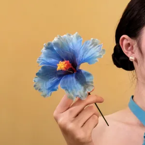 Close-up of a blue fabric hibiscus flower hair stick held by a hand against yellow