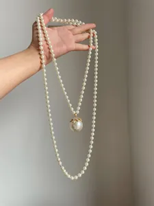 Dettaglio di una lunga collana di perle con grande pendente di perla e strass tenuta in mano.