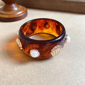 Primo piano di un bracciale in resina tartarugata con conchiglie dorate e perle bianche.