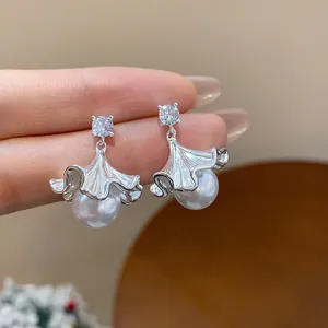 Detailed close-up of elegant zircon and silver ginkgo leaf pearl stud earrings between fingers.