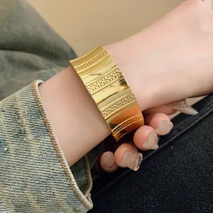 Detail of a wide gold-tone textured open cuff bracelet worn on the arm over denim.