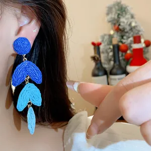 Close-up view of blue raffia woven tiered leaf drop earrings with silver needles on ear.