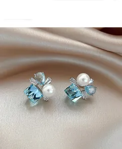 Macro detail of elegant square blue zircon and round pearl stud earrings on champagne silk
