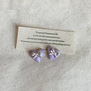 Detailaufnahme violetter Herzohrringe mit silberner Schleife auf cremefarbenem Stoff