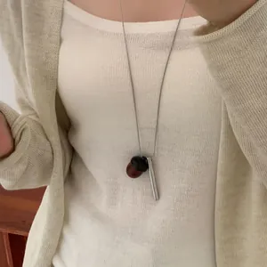Détail d'un long collier avec pendentifs gland et barre argentée sur un gilet beige.