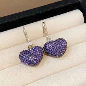 Elegant close-up of sparkly purple pavé crystal heart earrings in a beige jewelry box.