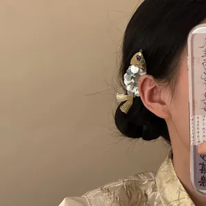 Détail d'une pince à cheveux poisson dorée avec écailles argentées sur des cheveux noirs.