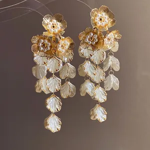 Boucles d'oreilles pendantes à fleurs dorées, perles et pétales blancs sur fond brun.