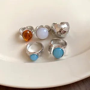 Detail showing five minimalist silver-tone open rings with various colorful stones in a dish.