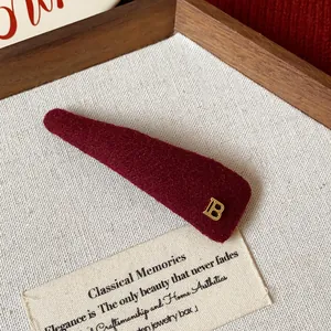 Close-up of a burgundy plush triangle hair clip with a golden 'B' letter detail.