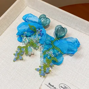 Close-up of blue chiffon flower earrings with green beaded heart and tassels on a jewelry tray.
