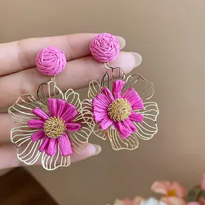 Nahaufnahme von pinken Raffia- und Goldblumenohrringen, die von Hand gehalten werden