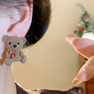 Close-up detail of a brown flocked teddy bear earring with a red bow worn on an ear.