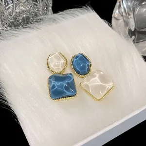 Pendientes cuadrados irregulares elegantes con textura brillante sobre fondo de piel blanca.