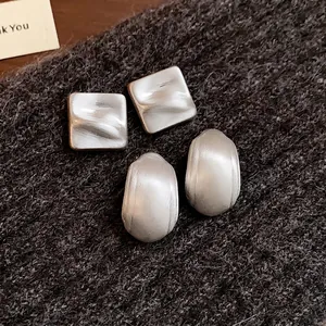 Orecchini a lobo minimalisti quadrati e ovali in argento opaco su sfondo di tessuto.
