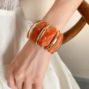 Nahaufnahme eines klobigen orangefarbenen Acryl- und Gold-Elastikarmbands am Handgelenk.