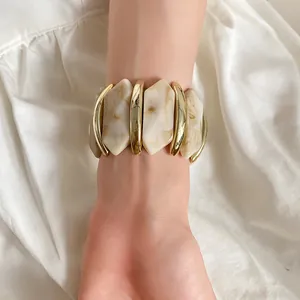 Detailansicht eines klobigen Armbands aus cremefarbenem Acryl und goldfarbenem Metall.