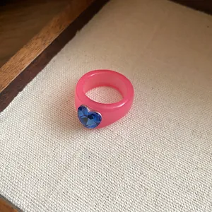 A close-up shot of a pink resin ring with a faceted blue heart-shaped rhinestone center.