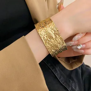 Close-up of a wide textured gold metal cuff bracelet on a female wrist in beige blazer