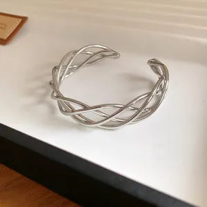 Primo piano di un bracciale rigido aperto intrecciato argento su un vassoio bianco minimalista.