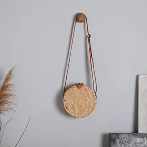 Détail d'un sac à bandoulière rond en paille tressée suspendu à un mur gris épuré.