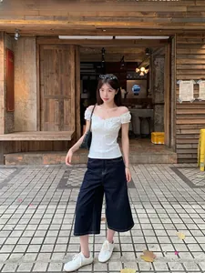 Full body shot of a woman in a white floral off-the-shoulder top and dark denim pants.