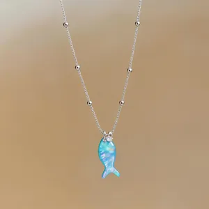 Close-up of a delicate silver beaded necklace featuring a vibrant blue opal fish pendant.