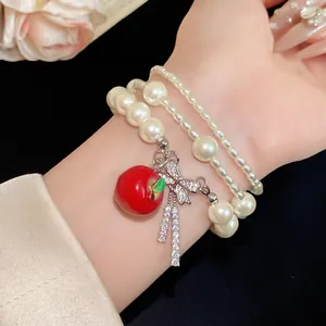 Detalle de una pulsera de perlas multicapa con dijes de manzana roja y lazo de circón.