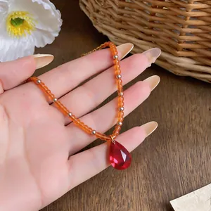 Détail d'un collier avec pendentif goutte rouge et perles orange sur fond naturel.