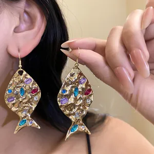Close-up of gold fish-shaped earrings with colorful rhinestones and hammered texture on model.