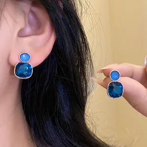 Elegant square blue gemstone earrings with silver pins shown on ear and held by hand