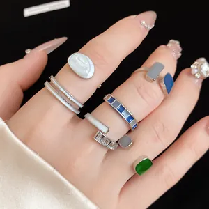 Dettaglio di vari anelli geometrici aperti in smalto blu e verde su mano curata.