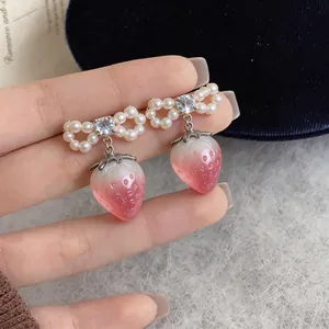 Close-up of elegant pearl bow earrings with pink strawberry pendants held by fingers.