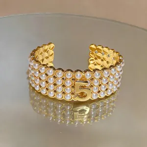 Elegante bracciale rigido aperto dorato con perle multistrato e un numero cinque centrale.