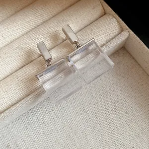 Close-up of silver-toned studs with transparent square resin pendants on cream jewelry box.