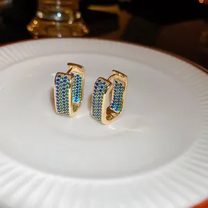 Close-up of gold-plated huggie earrings with blue and green pave zircons on a white plate.
