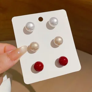 Close-up detail of three pairs of round pearl stud earrings in white, champagne, and red colors
