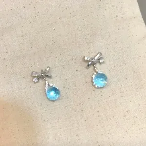 Silver bow stud earrings with light blue water-drop zircon pendants on beige background