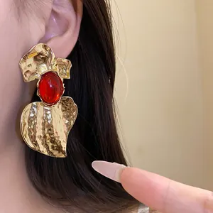 Close-up of oversized gold hammered heart earrings featuring a red resin stone inlay.