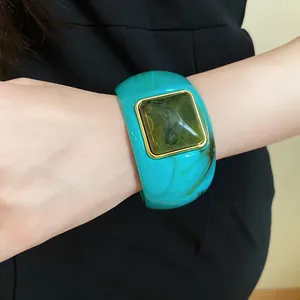 Close-up of a wide turquoise acrylic bangle with a square green resin centerpiece on a wrist.